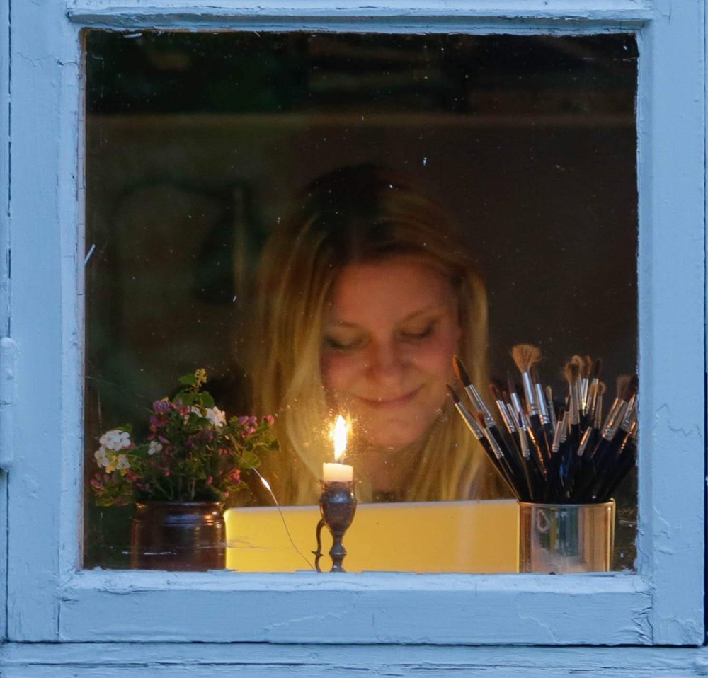 Sofie Bjarup bakom ett fönster, med ett tänt ljus, en blomvas och en burk penslar
